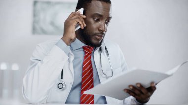 Afro-Amerikan doktor elinde bulanık dosya tutarken akıllı telefondan konuşuyor. 