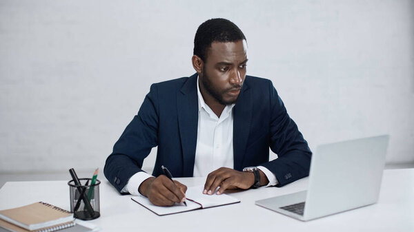 serious african american businessman writing on notebook while looking at laptop on grey 