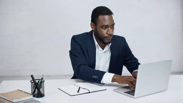 serious african american businessman typing on laptop near notebook on grey