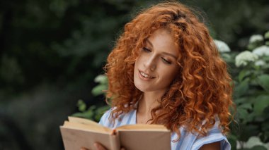Kıvırcık saçlı kadın dışarıda kitap okurken gülümsüyor.
