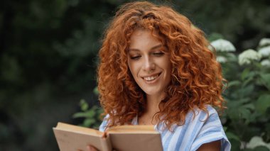 Dışarıda kitap okurken gülümseyen mutlu kızıl saçlı kadın. 