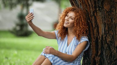 Mutlu kızıl kadın ağaç gövdesinin altında otururken selfie çekiyor. 