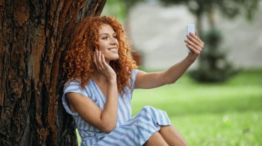 Mutlu kızıl saçlı kadın ağaç gövdesinin altında otururken selfie çekiyor. 