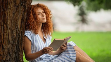 Çizgili elbiseli, elinde kitap olan ve ağaç gövdesinin altında oturan mutlu kızıl saçlı kadın. 