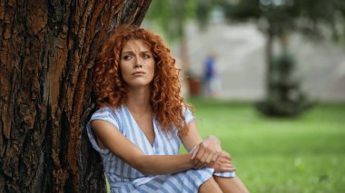 upset redhead woman in blue dress sitting under tree trunk in park clipart