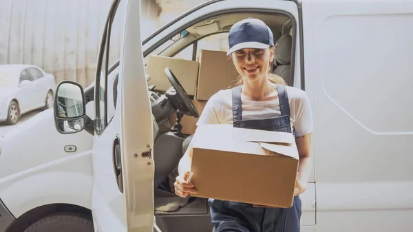 Smiling delivery woman holding carton box near car outdoors