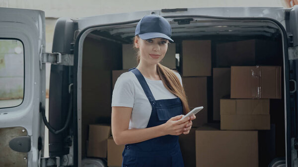 Delivery woman in overalls holding smartphone and looking at camera near cardboard boxes in car outdoors 