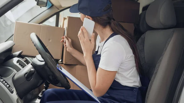 Delivery woman talking on cellphone near carton boxes and clipboard in car