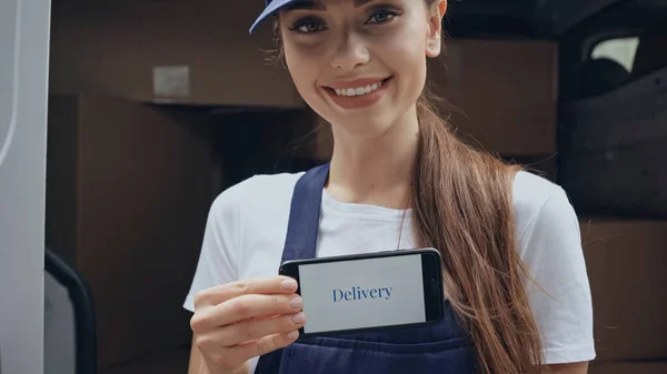 Smiling delivery woman holding mobile phone with delivery lettering near blurred car outdoors