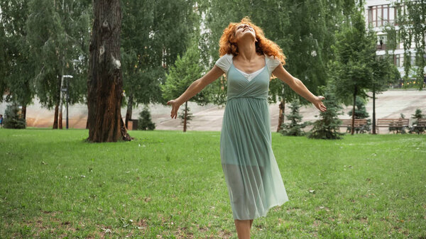 joyful redhead woman in chiffon dress standing in green park 