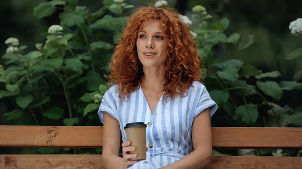 服を着た中赤頭の女が笑顔で紙コップを持ちながら公園のベンチに座って — ストック写真