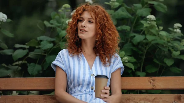 Encaracolado Ruiva Mulher Vestido Segurando Copo Papel Enquanto Sentado Banco — Fotografia de Stock