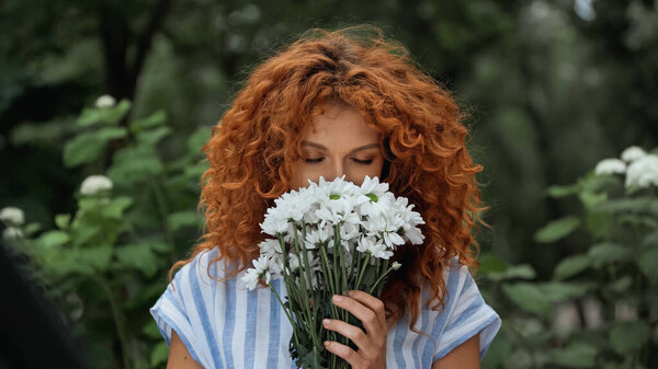 curly redhead woman with closed eyes smelling white flowers 