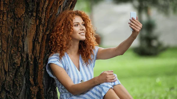 Alegre Pelirroja Mujer Tomando Selfie Mientras Sentado Bajo Árbol Tronco — Foto de Stock