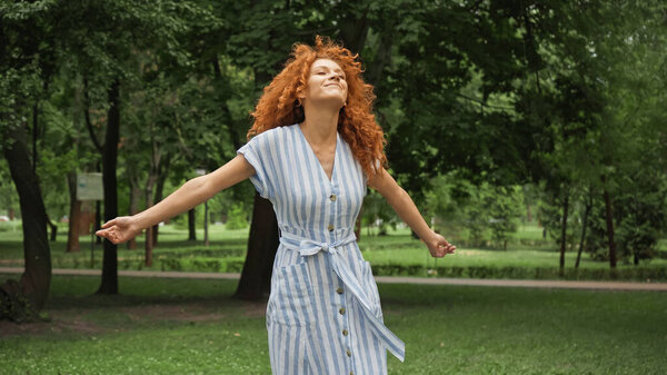 довольная девушка с рыжими волосами, стоящая с протянутыми руками в зеленом парке 