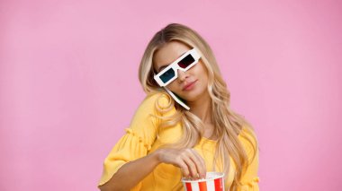 3D gözlüklü bir kadın akıllı telefonla konuşuyor ve pembe üzerinde patlamış mısır tutuyor. 