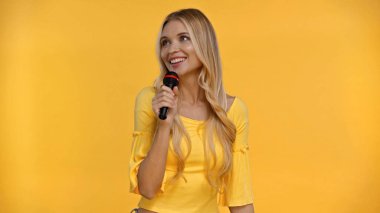 Positive blonde woman in blouse singing karaoke isolated on yellow  clipart