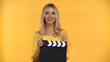 Happy blonde woman holding clapboard and looking at camera isolated on yellow  clipart