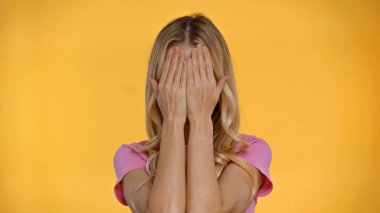 Blonde woman in t-shirt covering face with hands isolated on yellow  clipart