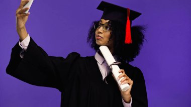 Gözlüklü ve mezuniyet şapkalı genç Afrikalı Amerikalı öğrenci mor renkte selfie çekiyor. 