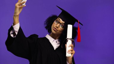 Gülümseyen Afro-Amerikan öğrenci mezuniyet şapkası ve cübbesi içinde mor üzerine izole edilmiş yuvarlanmış diplomasıyla selfie çekiyor. 