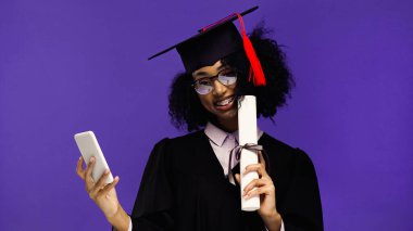Gülümseyen Afrikalı Amerikalı öğrenci mezuniyet şapkası ve cübbesi takmış elinde akıllı telefon ve diploması var. 