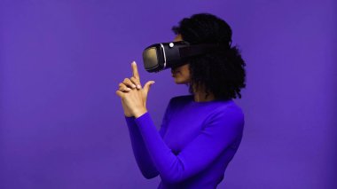 VR kulaklıklı kıvırcık Afrikalı Amerikalı kadın parmak tabancası jesti gösteriyor, elleri mor renkte.