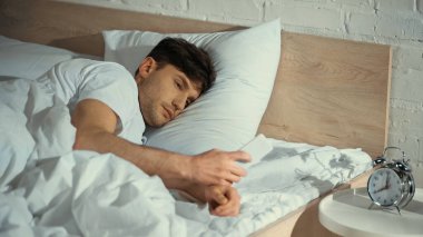 Adam yatakta yatarken akıllı telefona bakıyor. Komodinin yanında çalar saatin yanında.