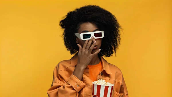 sad young african american woman in 3d glasses holding popcorn bucket while rubbing eye isolated on yellow