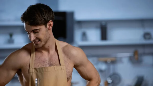 stock image shirtless man in apron smiling and looking down in kitchen