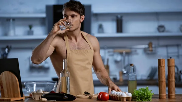 shirtless man in apron drinking white wine in modern kitchen