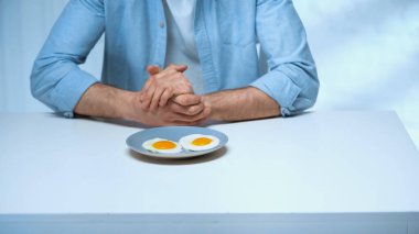 cropped view of man sitting with clenched hands near plate with fried eggs clipart