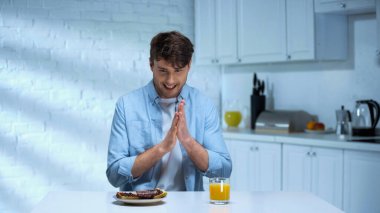 pleased man rubbing hands near toasts with confiture and orange juice in kitchen clipart