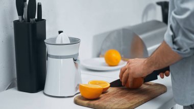 partial view of man cutting fresh orange near electric juicer in kitchen clipart