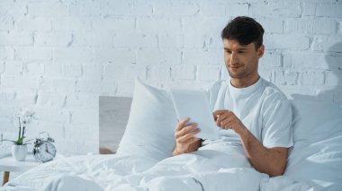 Yatakta yatarken komodinin yanındaki çalar saatin yanında dijital tablet kullanan gülümseyen adam.