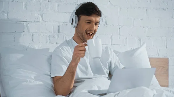 Laughing Man Headphones Holding Cup While Looking Laptop Bed — Stock Photo, Image