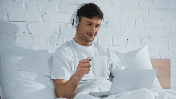 Sorrindo Homem Fones Ouvido Segurando Copo Usar Laptop Cama — Fotografia de Stock