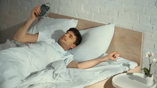 Sorrindo Homem Olhando Para Relógio Alarme Vintage Enquanto Deitado Cama — Fotografia de Stock