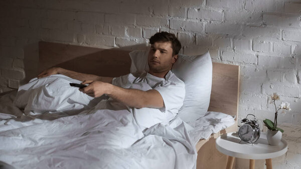 man clicking channels while watching tv in bed near alarm clock and orchid on bedside table