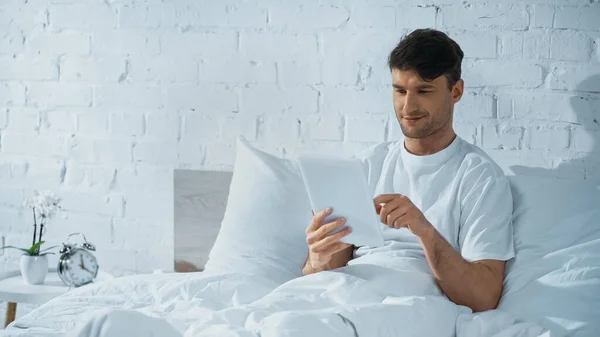 Mosolygós Férfi Használ Digitális Tabletta Miközben Ágyban Közelében Ébresztőóra Éjjeliszekrényen — Stock Fotó