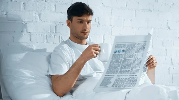 Człowiek Filiżanką Czytania Porannej Gazety Siedząc Rano Łóżku — Zdjęcie stockowe