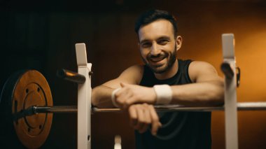 Koyu arkaplanda duran halterin yanındaki kameraya gülümseyen sporcu