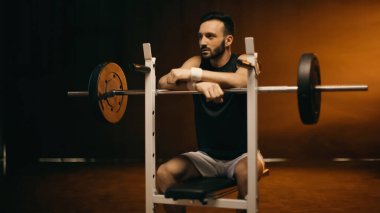 Sakallı sporcu, düz bankta, halterin yanında oturuyor.