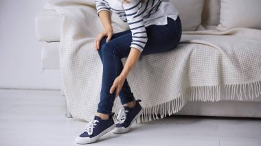 partial view of woman sitting on couch and touching painful leg clipart