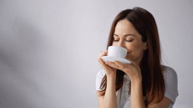 Gri arka planda sabah kahvesinin tadını çıkaran, kapalı gözlü mutlu bir kadın.