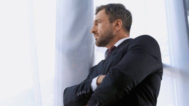 Siyah takım elbiseli işadamının düşük açılı görüntüsü pencerenin yanında çapraz kollarla duruyor.
