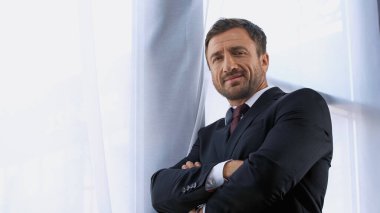 Ofiste kollarını kavuşturmuş kameraya bakan gülümseyen iş adamı.