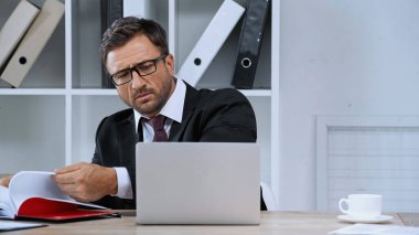 Gözlüklü ciddi bir işadamı ofiste dizüstü bilgisayarla çalışıyor.