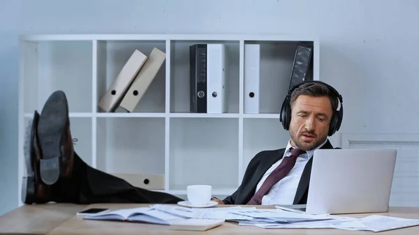 Geschäftsmann Mit Kopfhörern Sitzt Mit Beinen Auf Schreibtisch Neben Laptop — Stockfoto