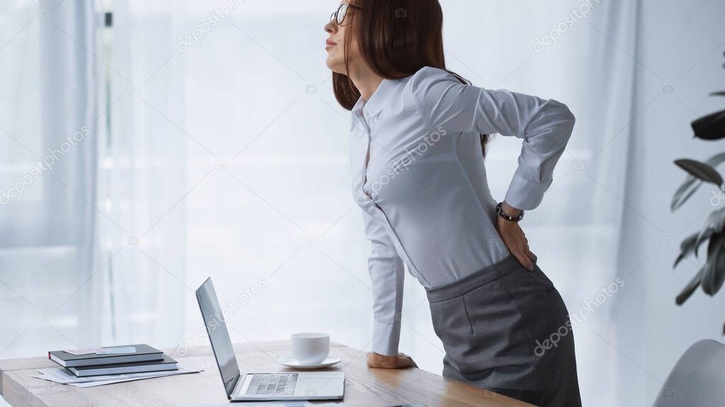 brunette woman standing at work desk and touching painful loin